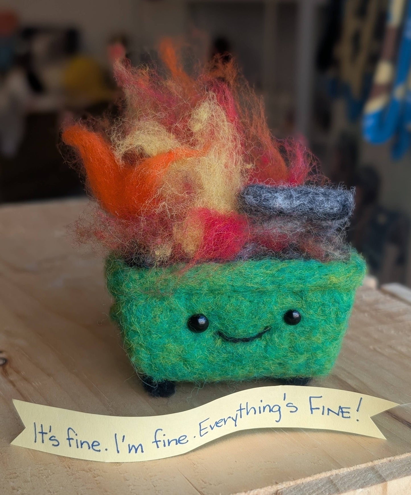 Colorful felted dumpster fire creature with a message ribbon that reads "it's fine. I'm Fine. Everything's fine!" on a wooden surface