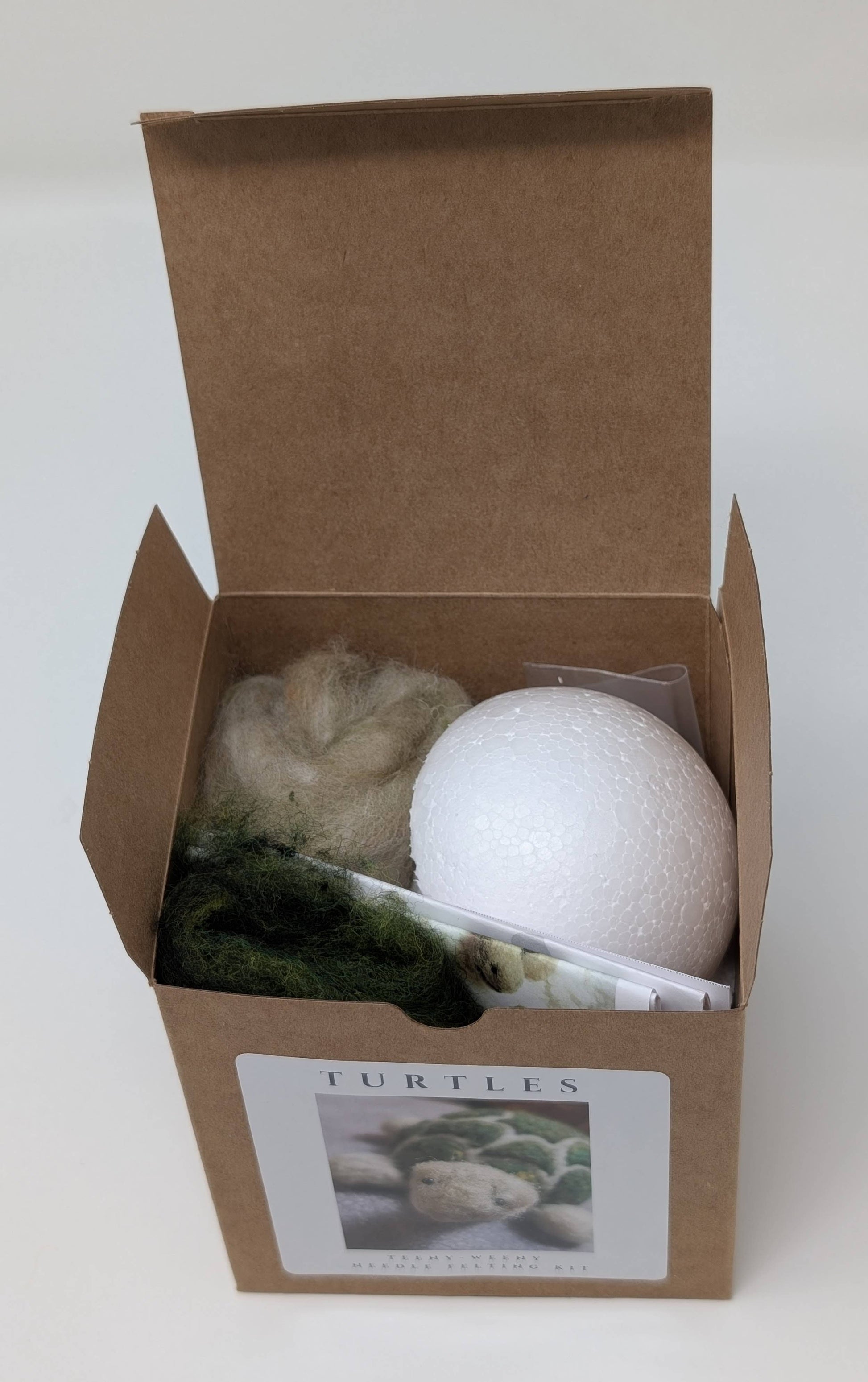 Open cardboard box containing needle felting related items on a white background